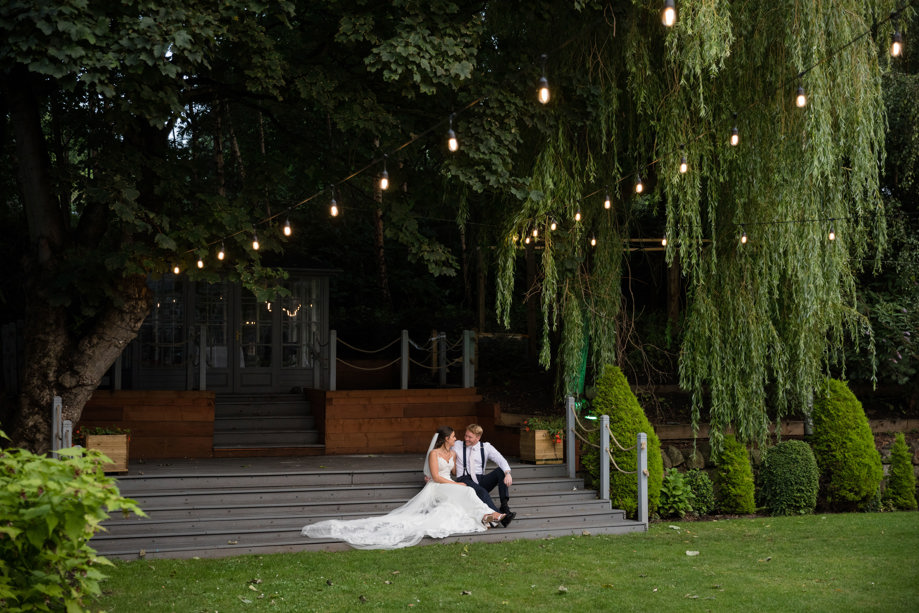 Bride and groom sit underneath festoon lights and hanging trees at The Venue, Eskmills